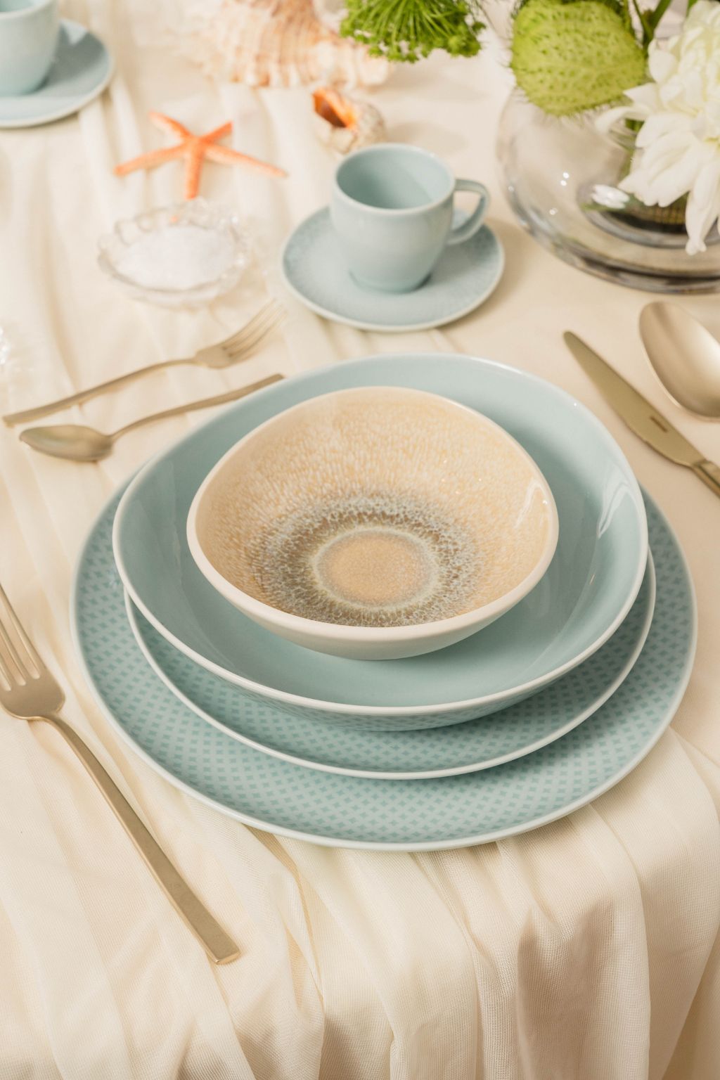 Stylishly set table with layered Rosenthal tableware in soft blue and beige tones, surrounded by elegant cutlery on a cream-colored tablecloth.