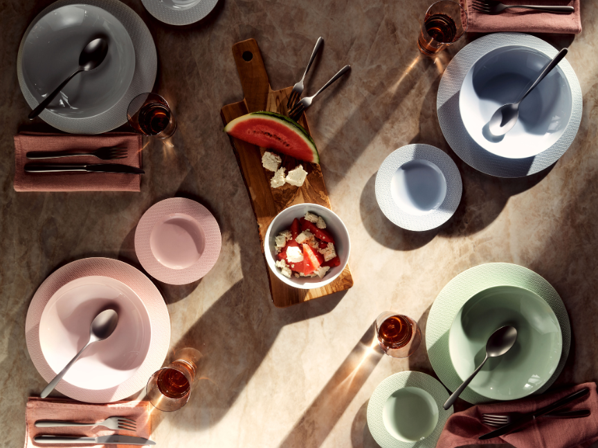 A top view of a stylishly laid table featuring Rosenthal tableware in delicate pastel shades such as pink, mint green and light blue. Plates, bowls and cups are arranged harmoniously and complemented by cutlery, cloth napkins and a serving platter with fresh fruit.