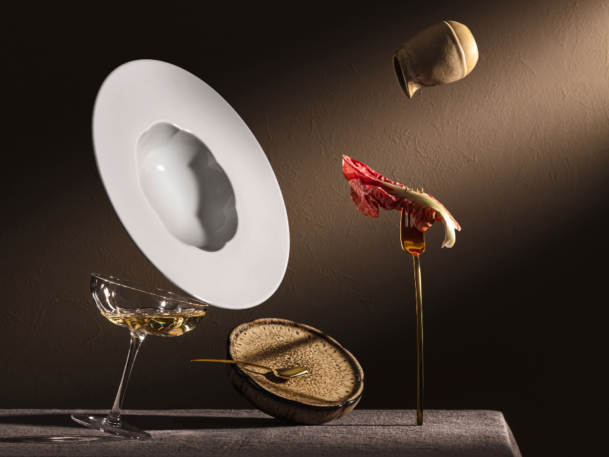 An artistically staged still life featuring Rosenthal porcelain and glass objects against a dark background. A white plate, elegant glasses and floating natural materials are dramatically lit, creating a modern composition.