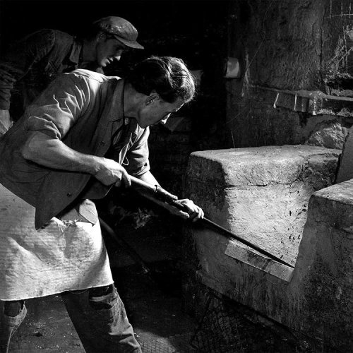 Worker engaged in traditional porcelain production, using a long tool to tend the kiln.