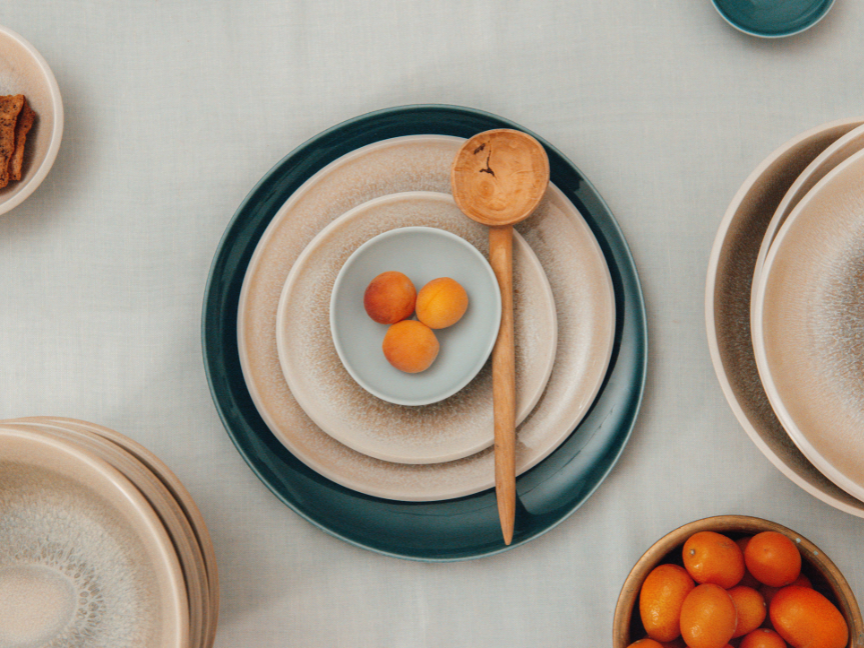 A top view of a modern arrangement of Rosenthal tableware in natural beige and petrol blue tones. Several plates with a textured surface are arranged in concentric circles.
