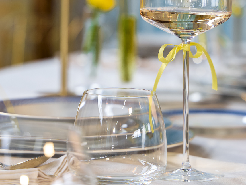 Close-up of an elegantly set table with Rosenthal glasses, white wine glass tied with a yellow ribbon, water glass, and delicate floral d&eacute;cor in the background