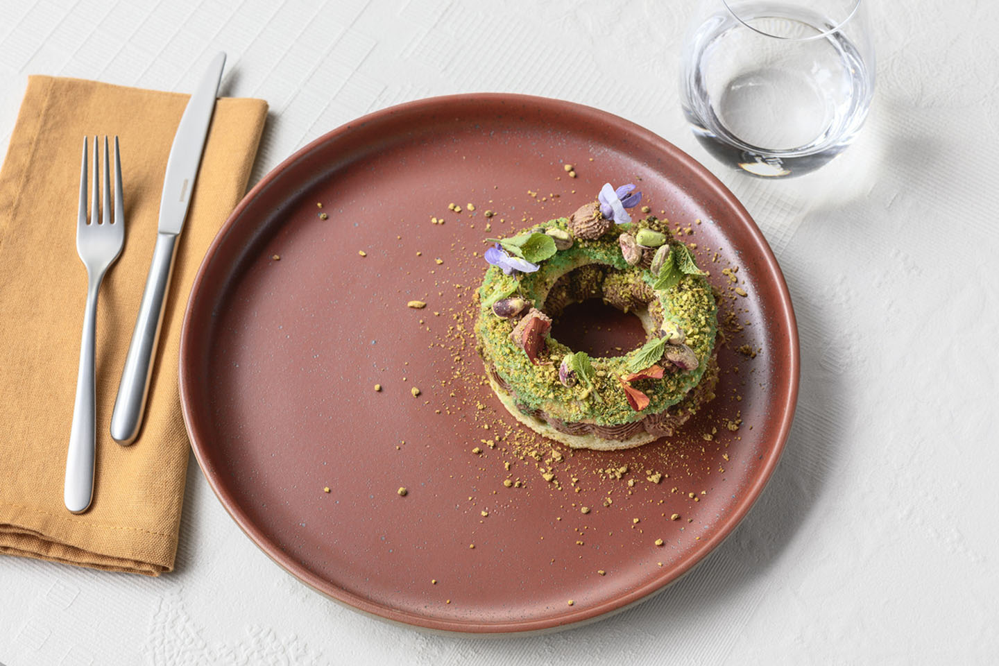 Rosenthal Geschirr in Mocha Mousse mit dekorativem Pistaziengeback, gelber Serviette, silbenem Besteck und einem Wasserglas.