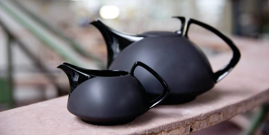 Two black TAC teapots stand one behind the other on a shelf