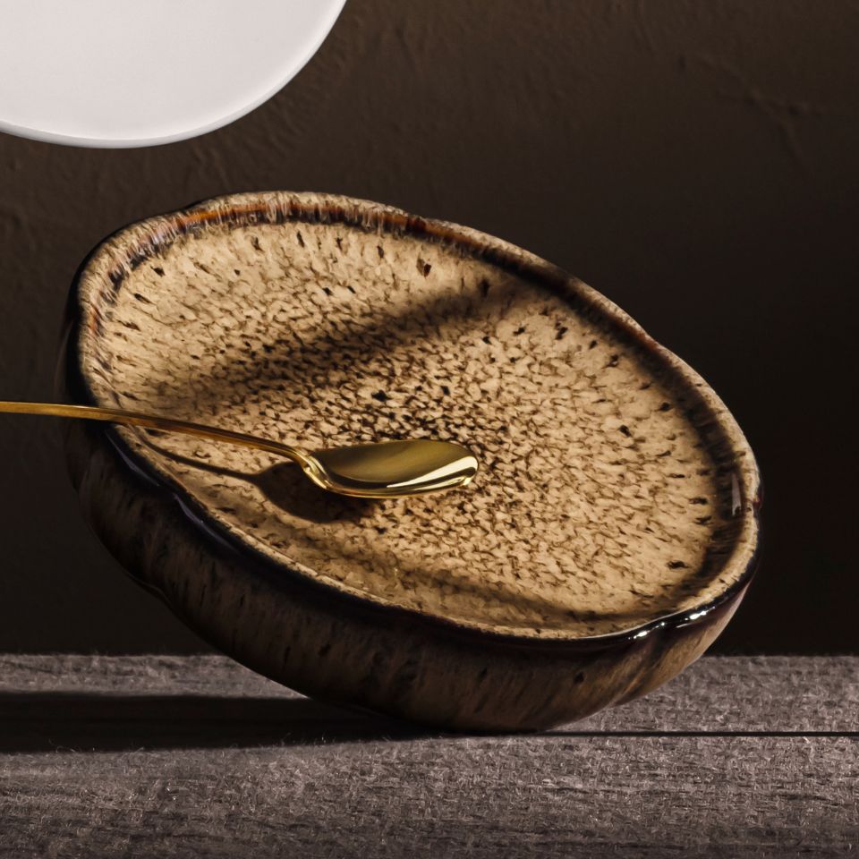 A close-up of a Rosenthal plate with a natural, speckled glaze in shades of beige and brown. A gold-coloured spoon rests on the plate, highlighting the high-quality materials and the modern design inspired by organic forms